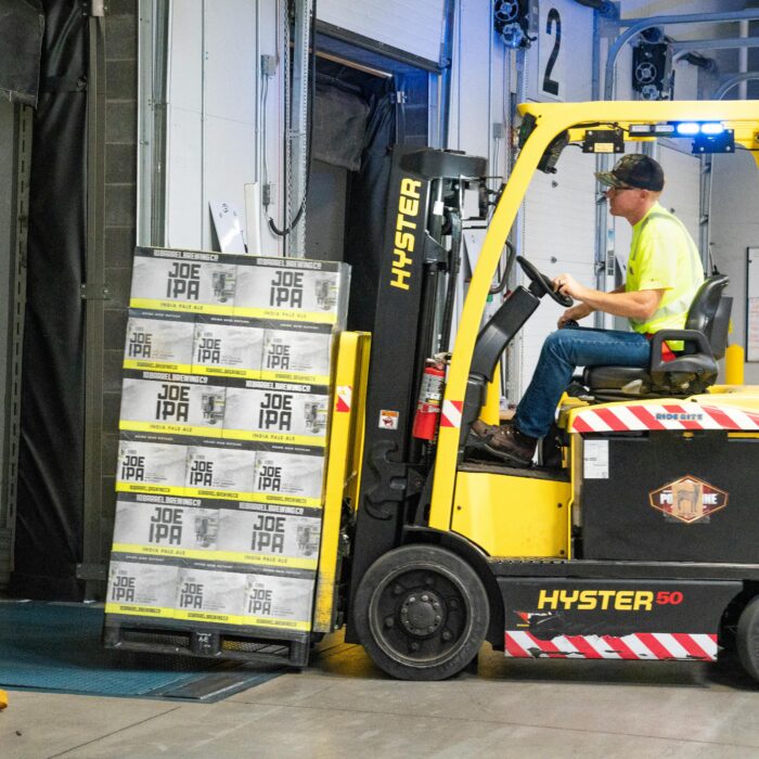 A worker drives a Hyster forklift moving Joe IPA boxes in a warehouse.